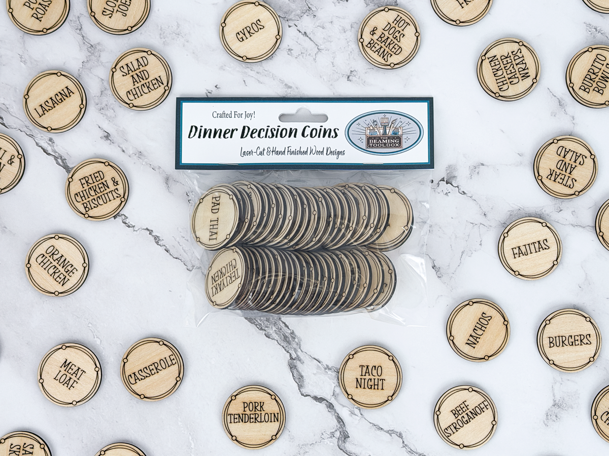 A package of laser-cut wooden dinner decision coins lies on a white marble surface, surrounded by loose matching coins engraved with food options like tacos, burgers, and pizza night. The package label reads 'Dinner Decision Coins' and shows the brand 'The Beaming Toolbox'.