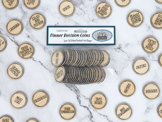 A package of laser-cut wooden dinner decision coins lies on a white marble surface, surrounded by loose matching coins engraved with food options like tacos, burgers, and pizza night. The package label reads 'Dinner Decision Coins' and shows the brand 'The Beaming Toolbox'.