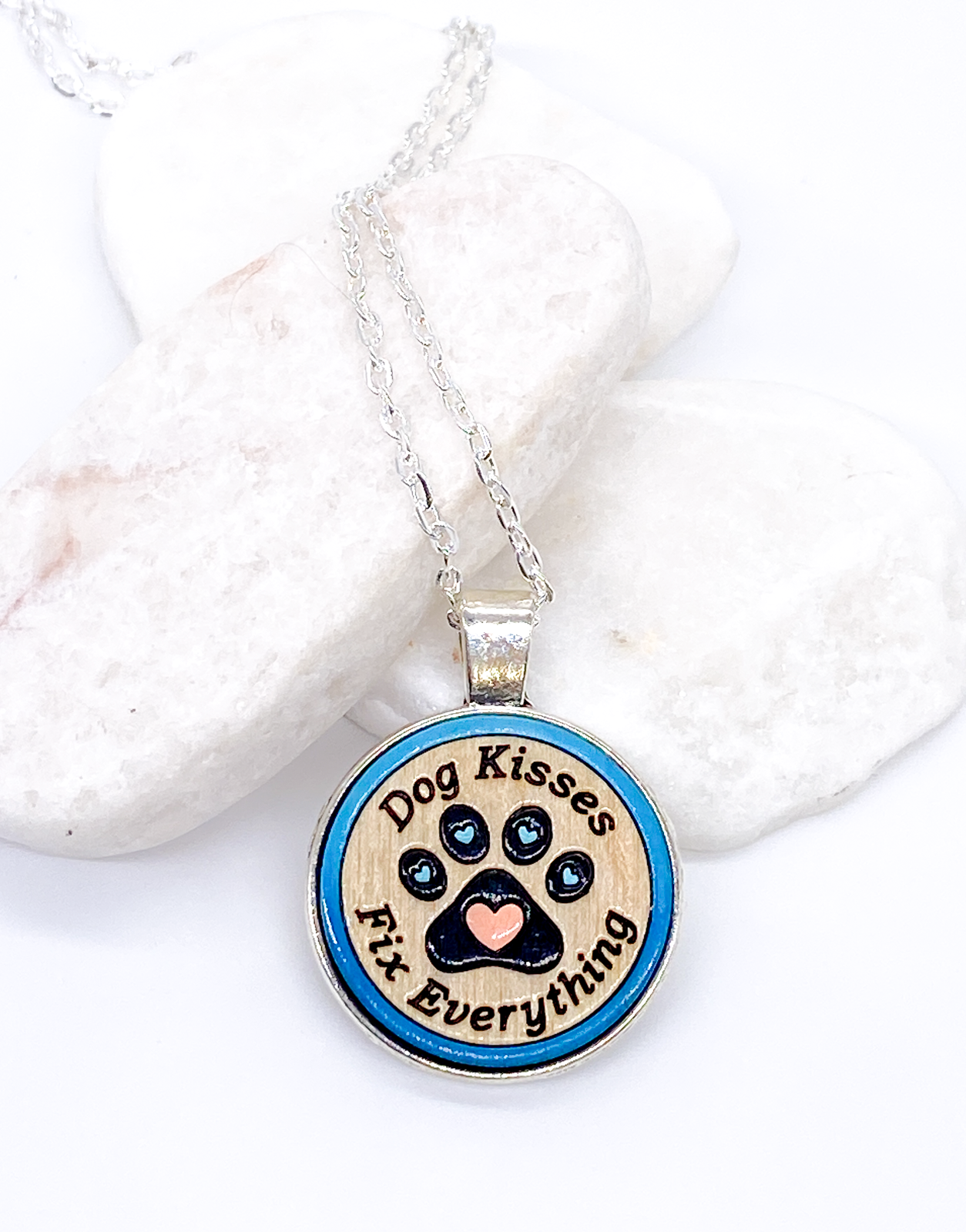 Dog kisses wood pendant necklace with blue painted paw print design displayed on white stones, flat lay on white background