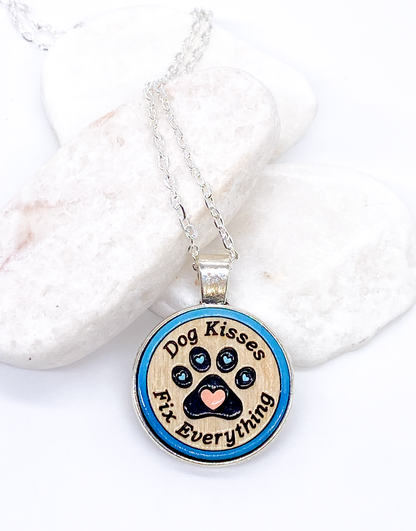 Dog kisses wood pendant necklace with blue painted paw print design displayed on white stones, flat lay on white background