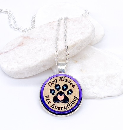 Dog kisses wood pendant necklace with purple painted paw print design displayed on white stones, flat lay on white background