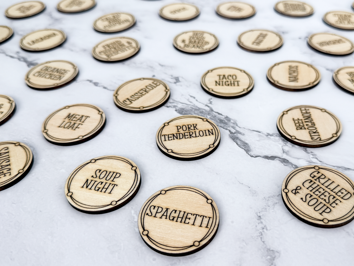 A collection of round, laser-cut wooden tokens engraved with various dinner options like 'Spaghetti', 'Meat Leaf', and 'Soup Night' arranged on a grey marble surface.