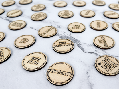 A collection of round, laser-cut wooden tokens engraved with various dinner options like 'Spaghetti', 'Meat Leaf', and 'Soup Night' arranged on a grey marble surface.
