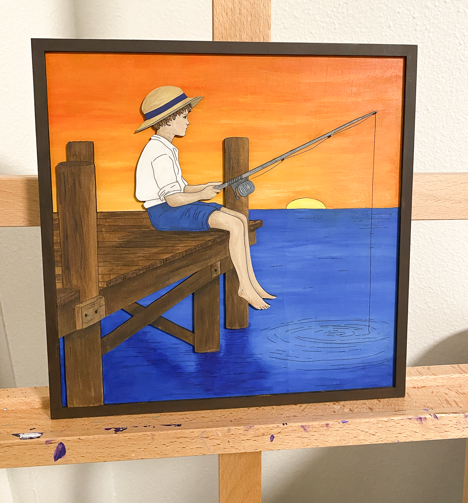 Painted layered wood fishing boy art showing child sitting on dock over blue water with yellow and orange sunset sky sitting on a painting easel