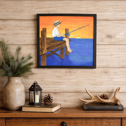 Painted layered wood diy art kit of a  child sitting on dock over blue water with yellow and orange sunset sky  displayed on a wooden wall above a wooden console table.