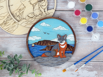 Finished island fox adult craft kit shown with painted layered wood artwork, unpainted kit pieces, paint pots, and brushes on a rustic wood background.