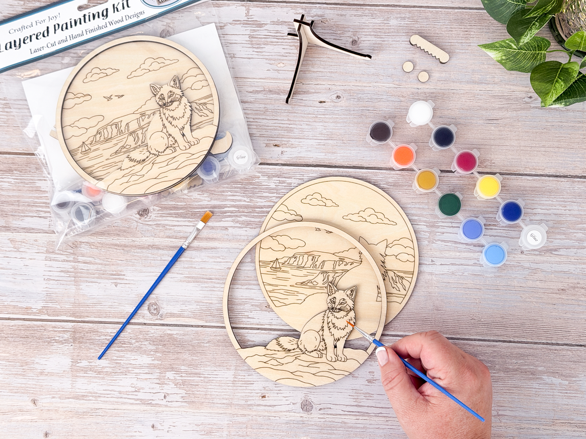Person painting the Fox Coastal layered wood craft kit on a rustic background with paint pots, brushes, packaging, and display hardware.
