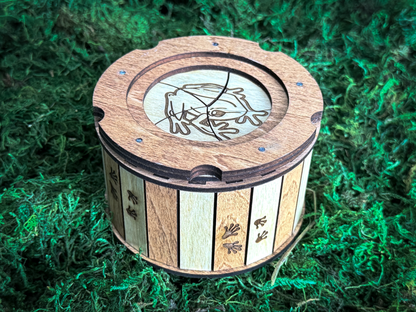 Close-up of a wooden frog iris keepsake box with an engraved frog on the lid, alternating natural and stained wood panels, and tiny frog footprints.
