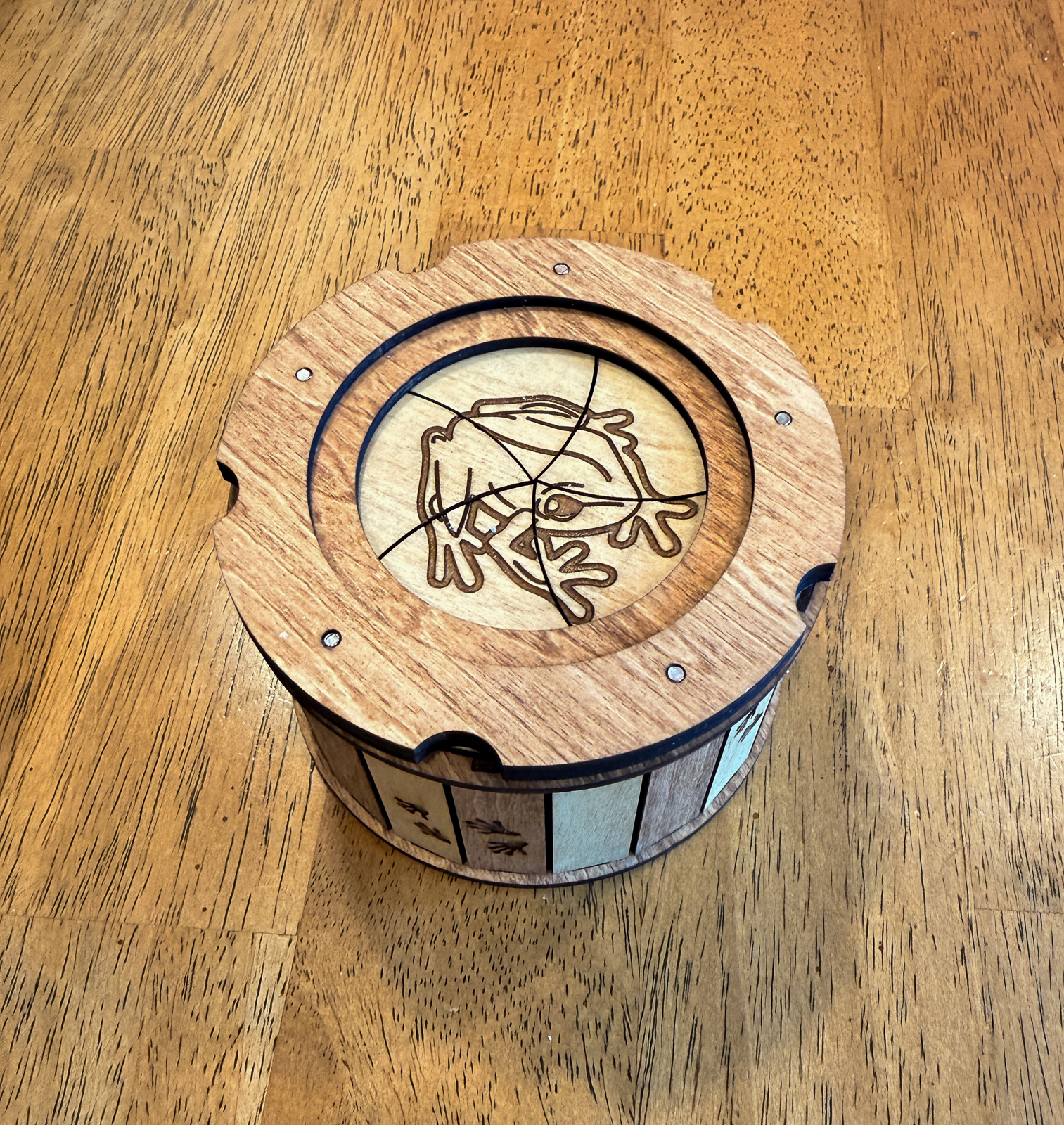Handcrafted wooden Frog Keepsake Box with engraved frog lid and twist-open iris mechanism resting on a wood table.