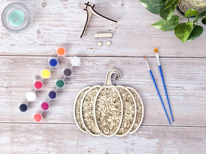 Person painting the Gothic Pumpkin layered wood craft kit on a rustic background with paint pots, brushes, packaging, and display hardware.