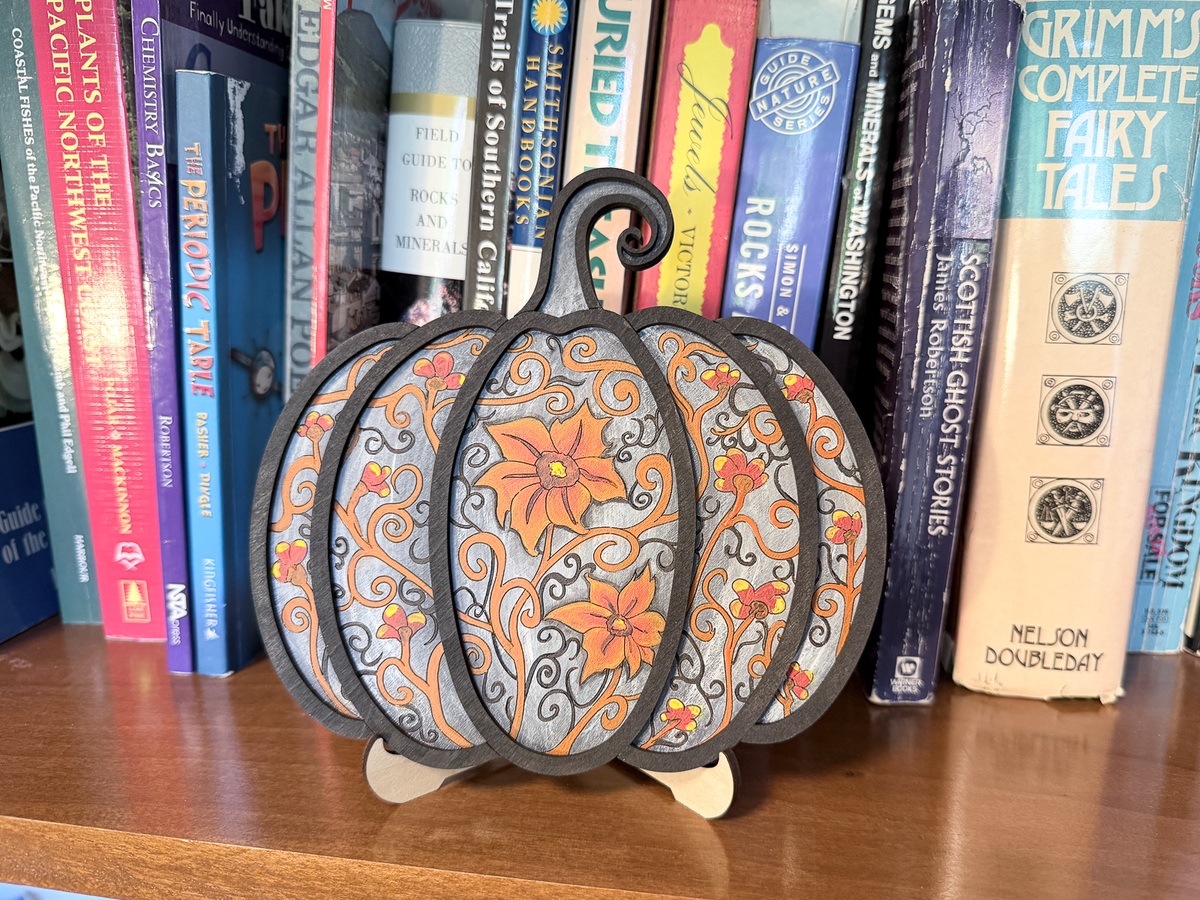 Finished Gothic Pumpkin layered wood artwork painted in gray, orange, red, yellow, and black, displayed on a stand in front of books.
