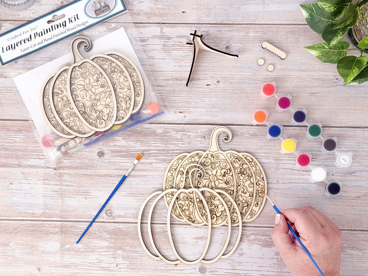 Person painting the Gothic Pumpkin layered wood craft kit on a rustic background with paint pots, brushes, packaging, and display hardware.