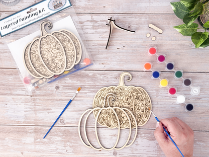 Person painting the Gothic Pumpkin layered wood craft kit on a rustic background with paint pots, brushes, packaging, and display hardware.