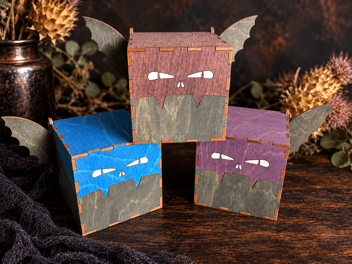 Three handcrafted wooden bat boxes in blue, brown, and purple stains with engraved white eyes, dark bat wings, and square wooden bodies on a rustic tabletop.