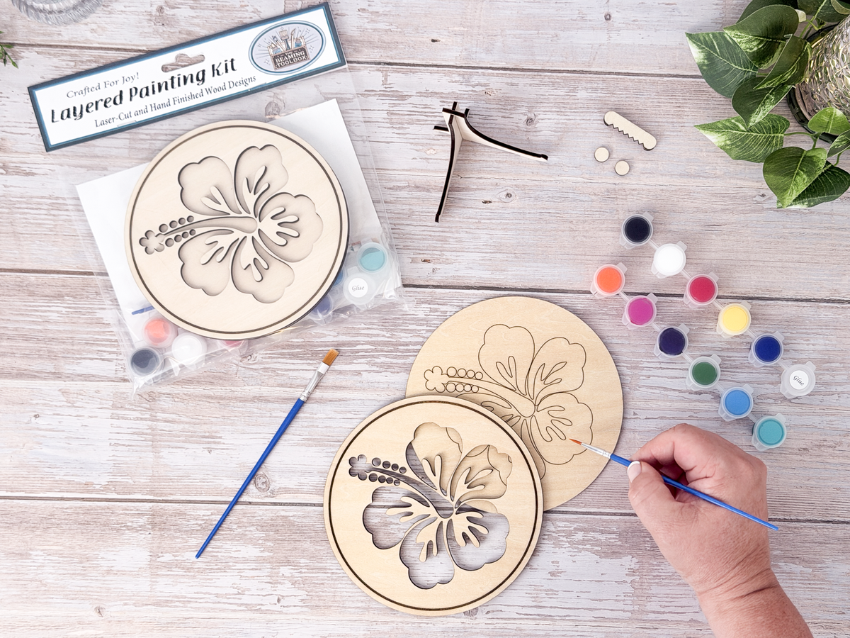 Person painting the Hawaiian Hibiscus layered wood craft kit with paint pots, brushes, stand, hardware, and packaging nearby.