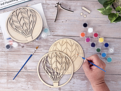Person painting the Hot Air Balloon layered wood craft kit with paint pots, brushes, stand, hardware, and packaging nearby.