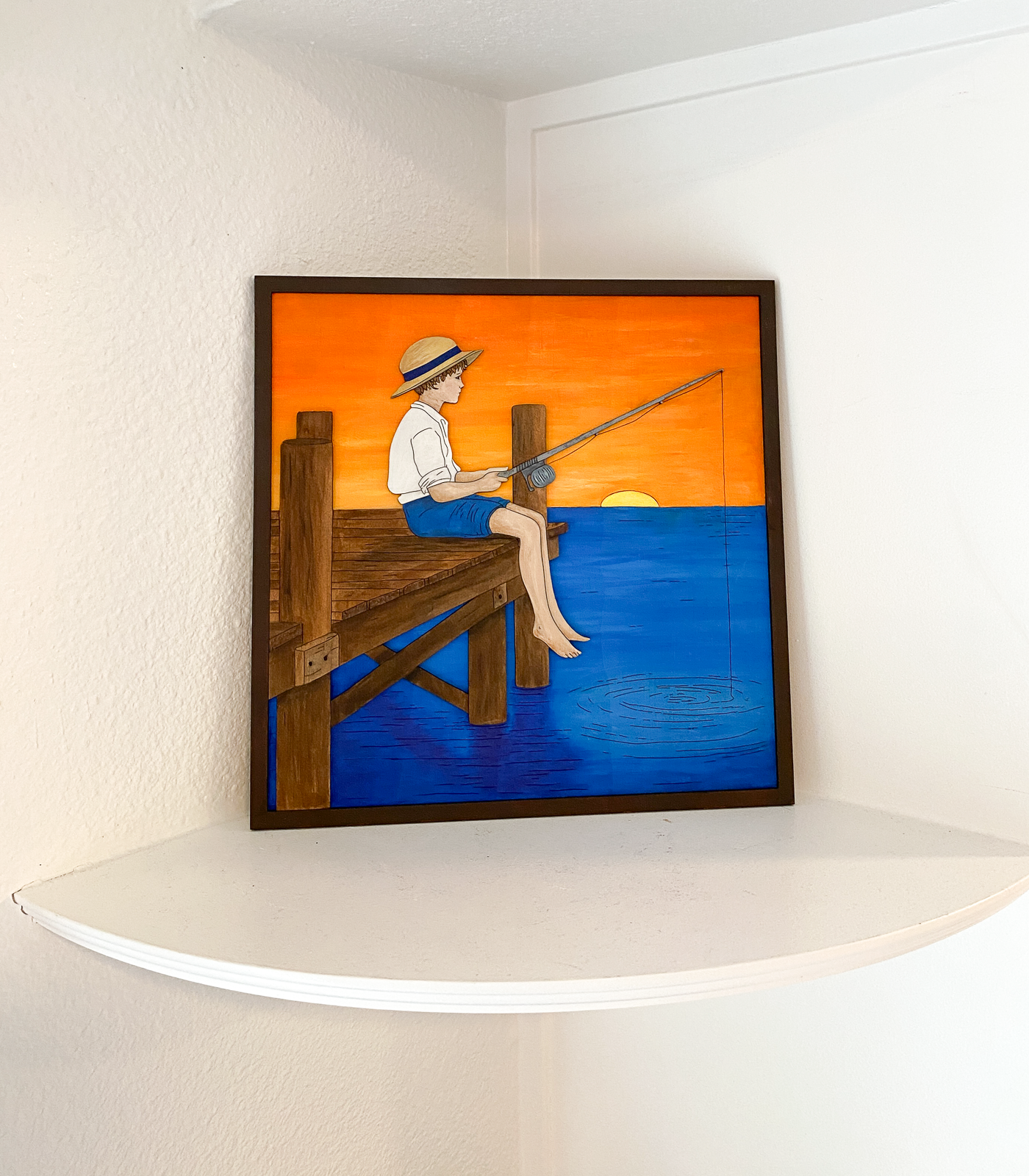 Framed artwork of a person fishing on a dock with an orange and blue background.