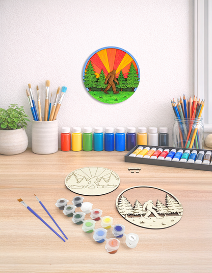 A finished Sasquatch wood kit displayed on a white wall in a bright studio workspace alongside the unpainted kit components and colorful art supplies