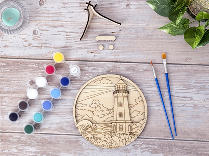 Unpainted lighthouse coastal layered wood artwork displayed with paint pots, brushes, stand pieces, candle, and greenery.