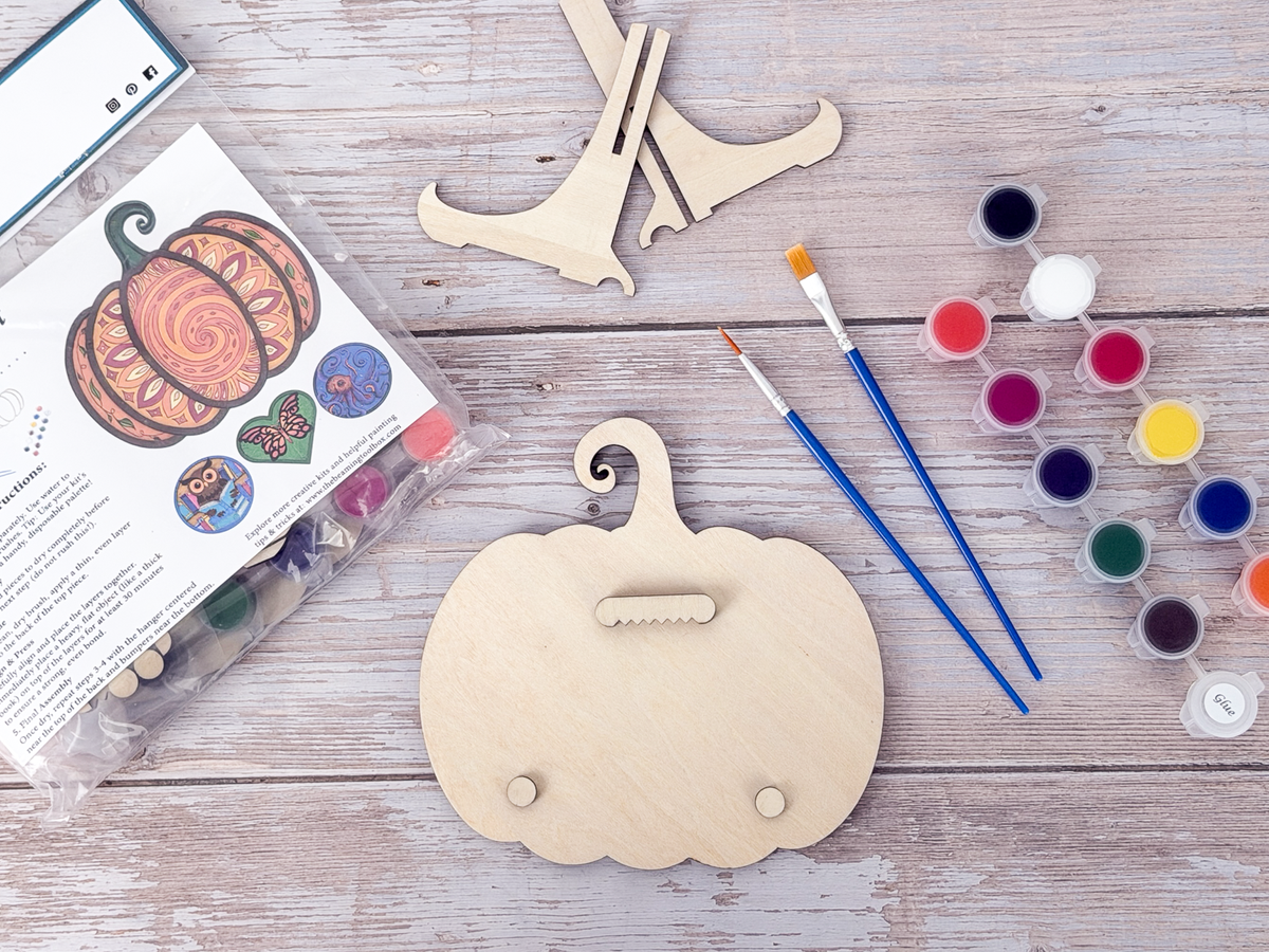 Back of the Mandala Pumpkin wood craft kit showing the hanger piece, small bumpers, display stand, paint pots, brushes, and packaging.