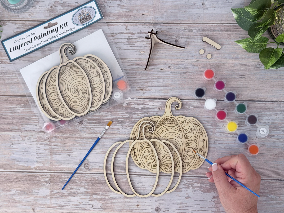Person painting the Mandala Pumpkin layered wood craft kit on a rustic background with paint pots, brushes, packaging, and display hardware.