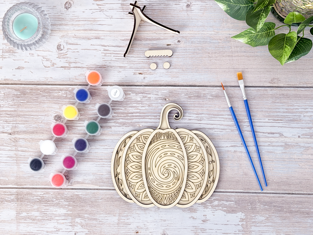 Unpainted Mandala Pumpkin layered wood artwork displayed with paint pots, brushes, stand pieces, candle, and greenery on a rustic background.