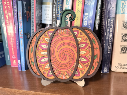 Finished Mandala Pumpkin layered wood artwork painted in orange, pink, yellow, green, and brown, displayed on a stand in front of books.