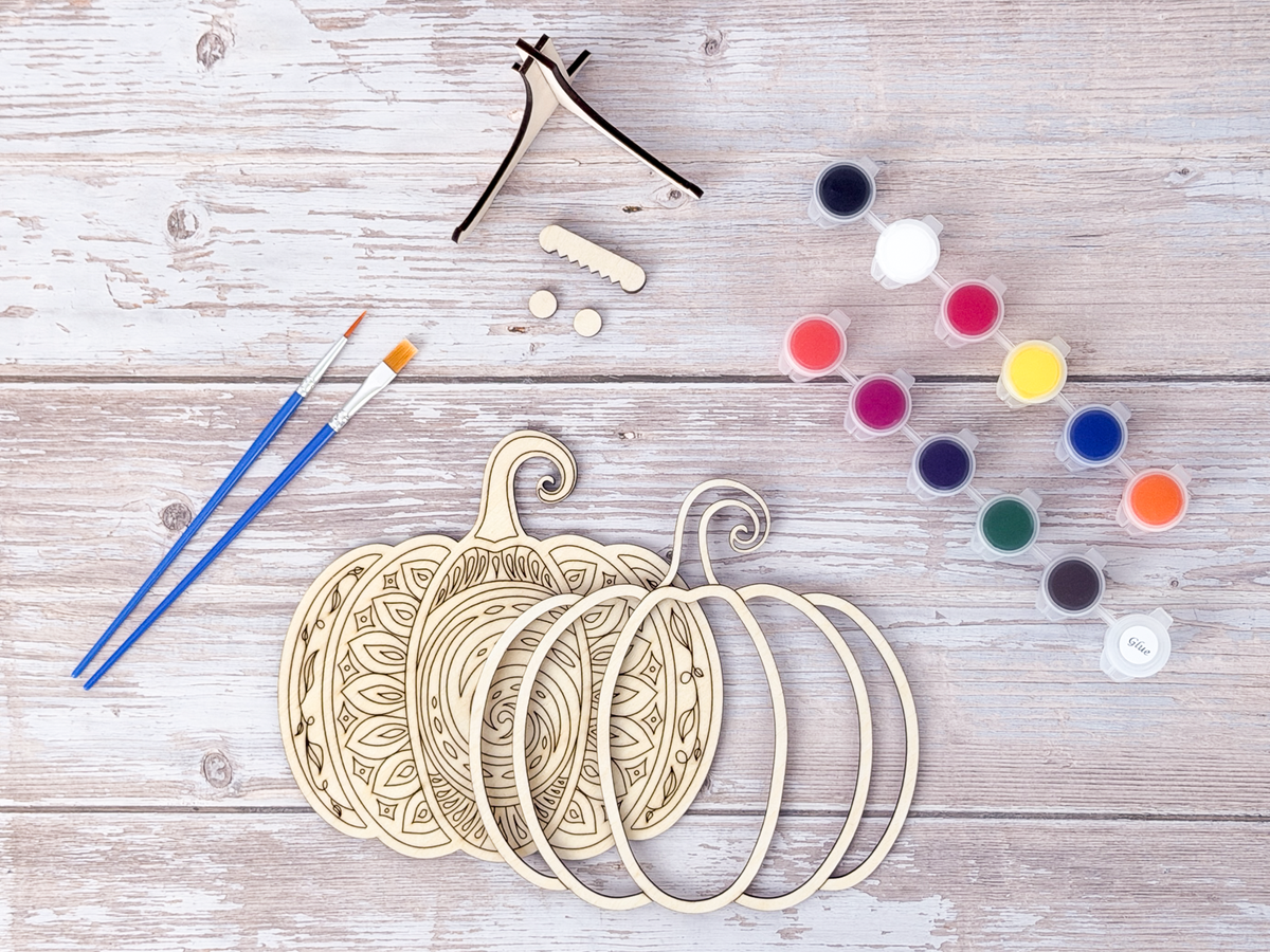 Unfinished Mandala Pumpkin wood craft kit pieces arranged with paint pots, brushes, display stand, hanger hardware, and bumpers.