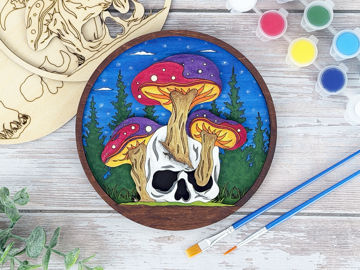 Finished mushroom skull adult craft kit shown with painted layered wood artwork, unpainted kit pieces, paint pots, and brushes on a rustic wood background.