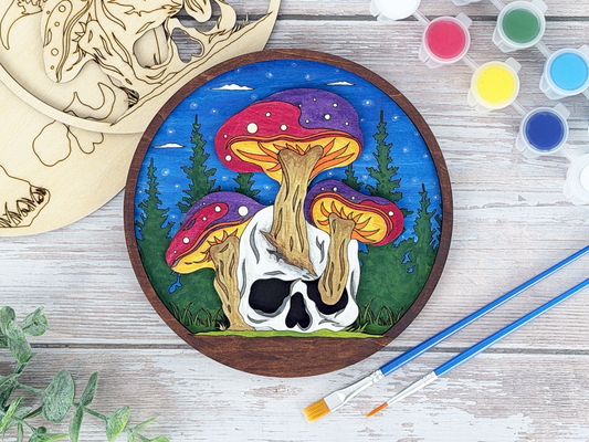 Finished mushroom skull adult craft kit shown with painted layered wood artwork, unpainted kit pieces, paint pots, and brushes on a rustic wood background.