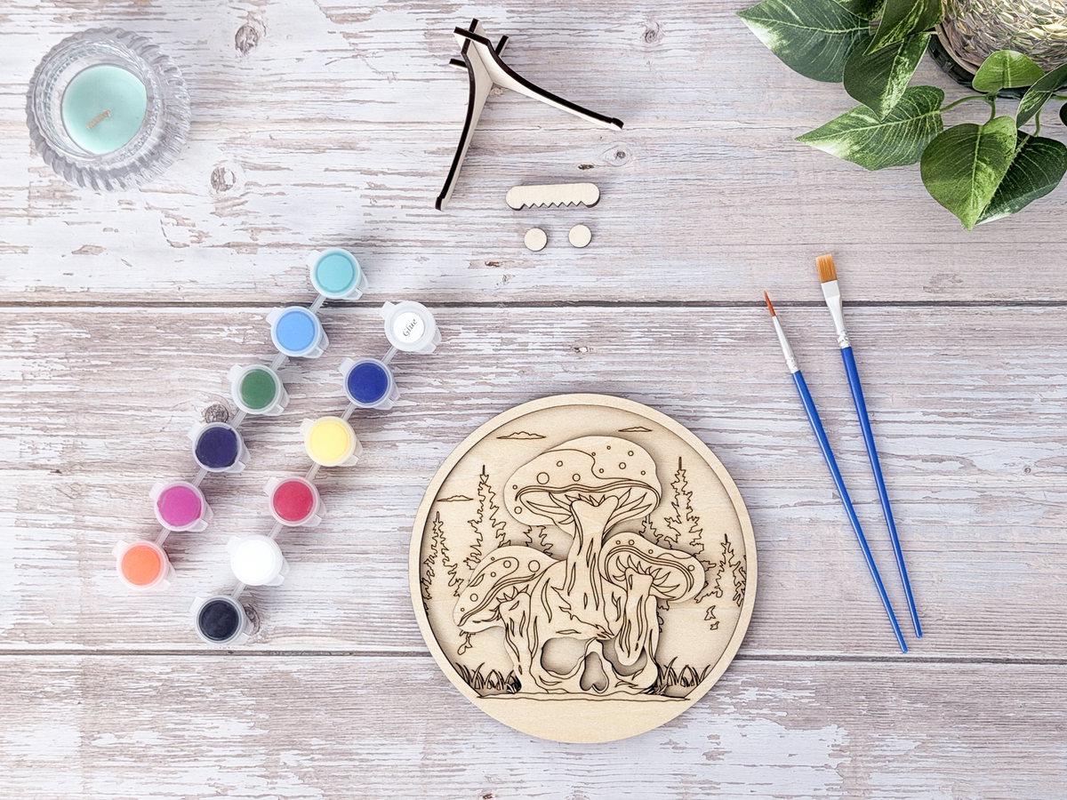 Unfinished Mushroom Skull layered wood artwork displayed with paint pots, brushes, stand pieces, candle, and greenery on a rustic background.