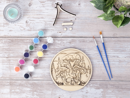 Unfinished Mushroom Skull layered wood artwork displayed with paint pots, brushes, stand pieces, candle, and greenery on a rustic background.