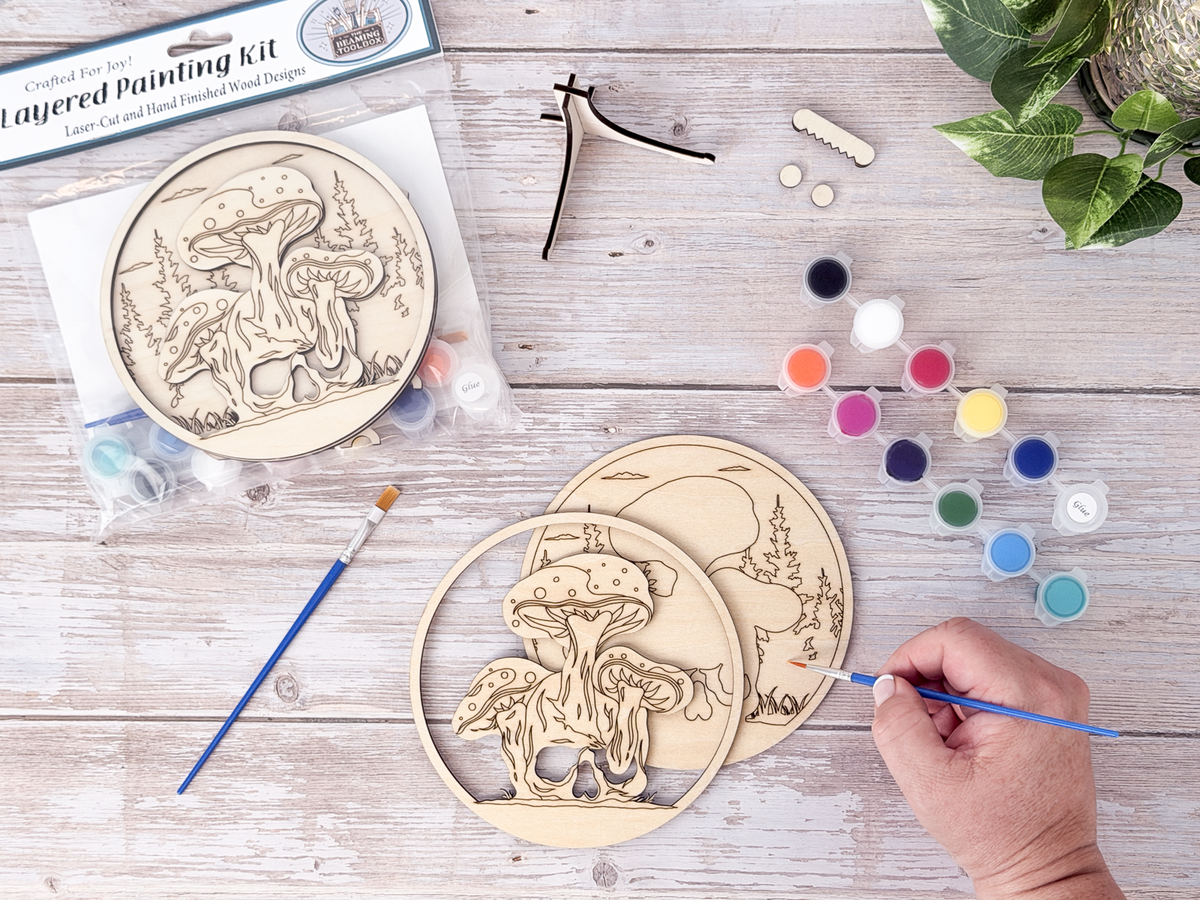 Person painting the Mushroom Skull layered wood craft kit on a rustic background with paint pots, brushes, packaging, and display hardware.