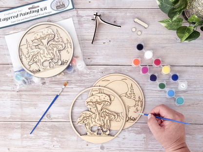 Person painting the Mushroom Skull layered wood craft kit on a rustic background with paint pots, brushes, packaging, and display hardware.