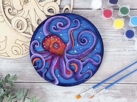 Finished octopus adult craft kit shown with painted layered wood artwork, unpainted kit pieces, paint pots, and brushes on a rustic wood background.