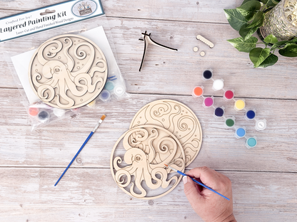 Person painting the octopus layered wood craft kit on a rustic wood background with paint pots, brushes, packaging, and display hardware.