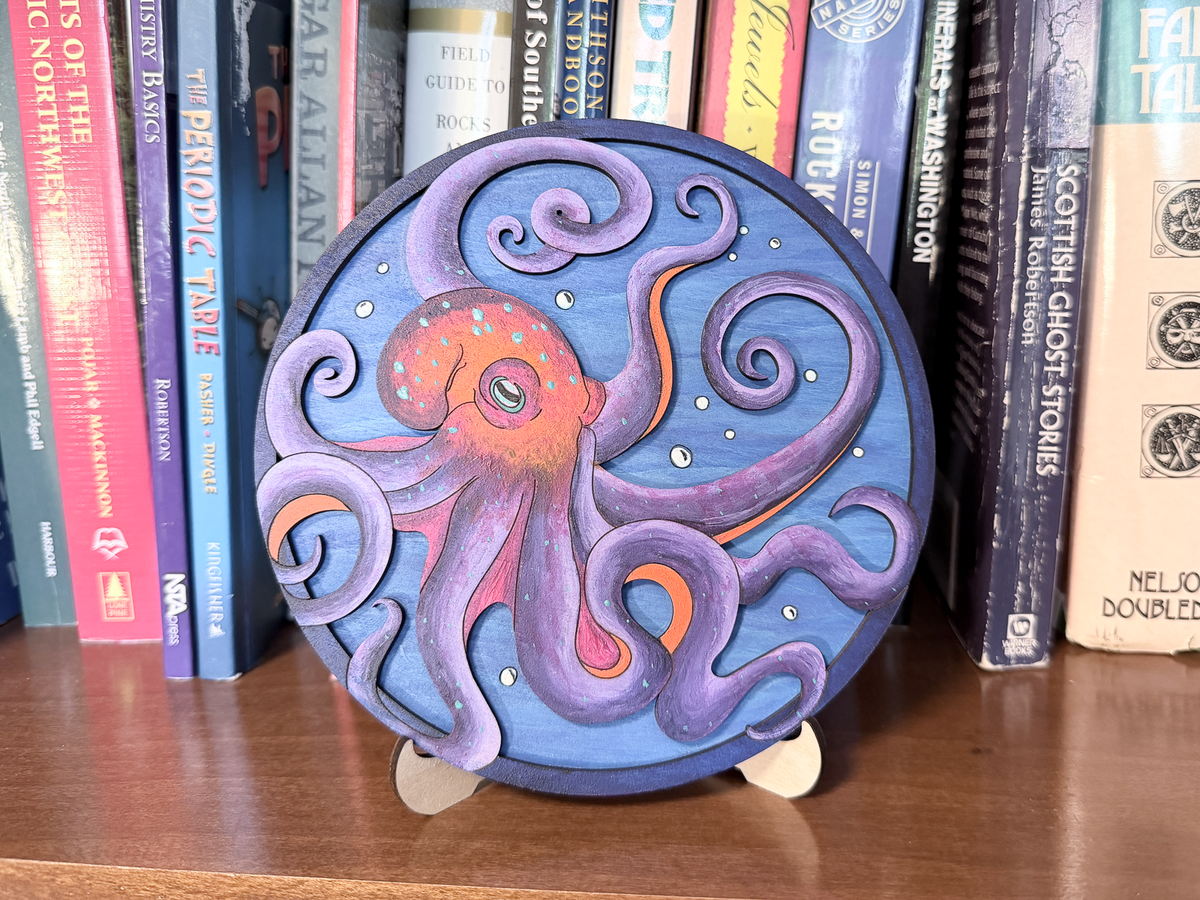 Finished octopus layered wood artwork painted in blue, purple, red, and orange, displayed on a stand in front of books.