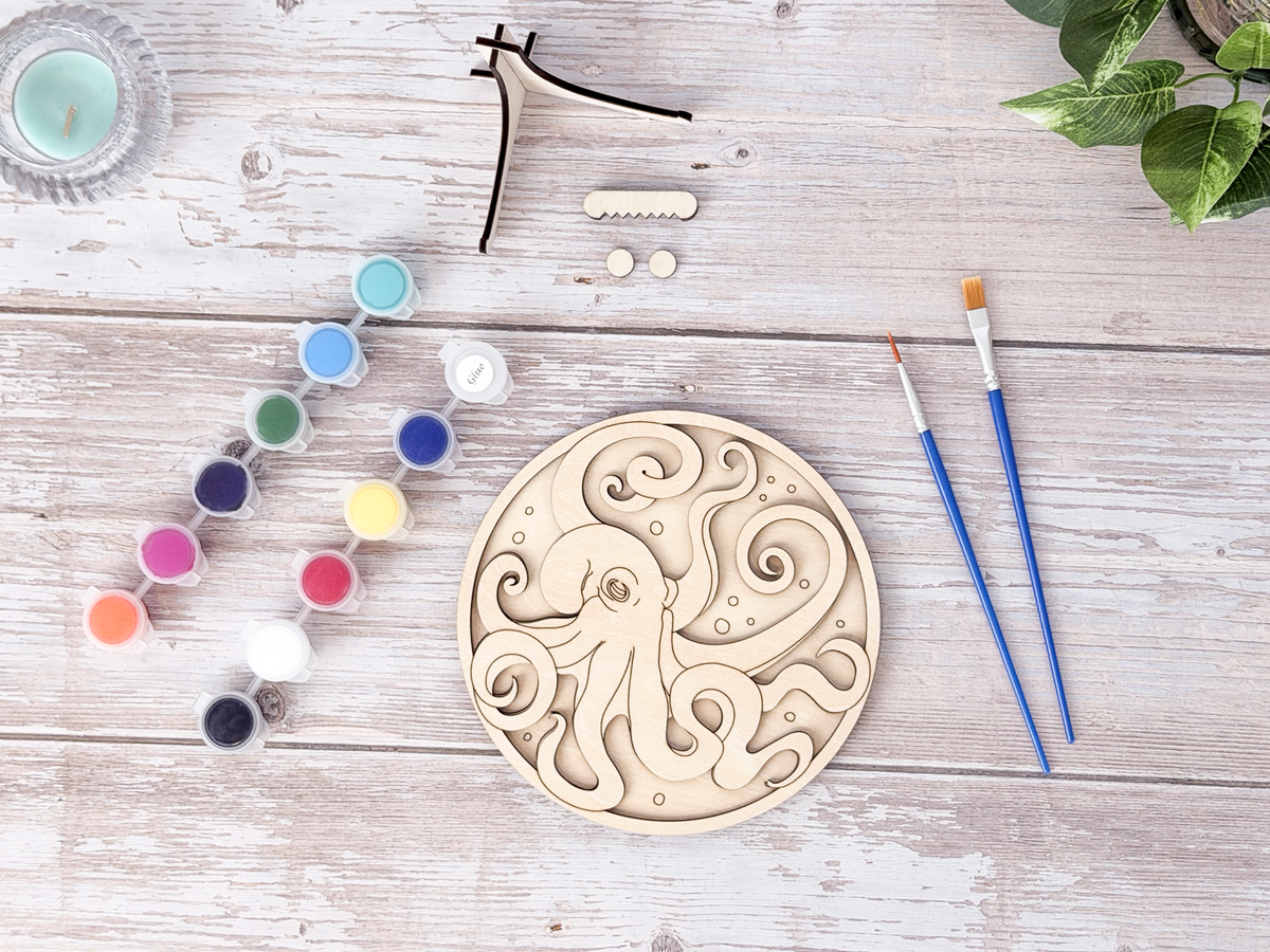 Unpainted octopus layered wood artwork displayed with paint pots, brushes, stand pieces, candle, and greenery on a rustic background.