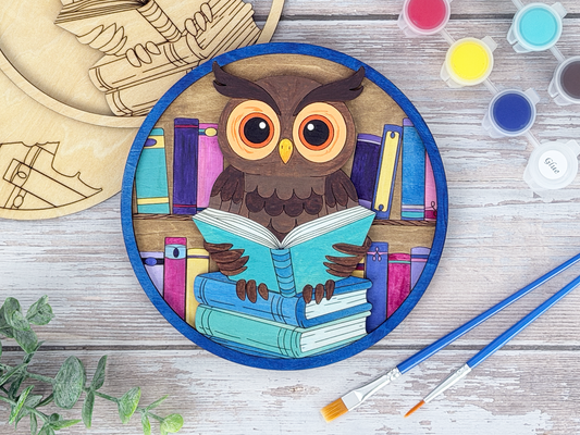 Finished book lovers owl adult craft kit shown with painted layered wood artwork, unpainted kit pieces, paint pots, and brushes on a rustic wood background.