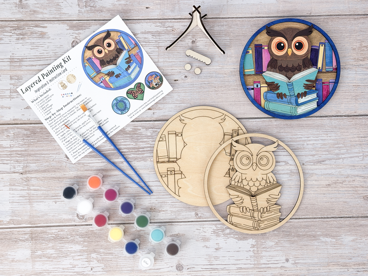 A layered painting kit featuring a circular wooden owl design, two paintbrushes, twelve small paint pots, a wooden display stand, and an instruction card titled Layered Painting Kit with step-by-step instructions and illustrations.