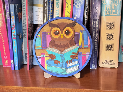 Finished book lovers owl layered wood artwork painted green, pink, orange, and black, displayed on a stand in front of books.