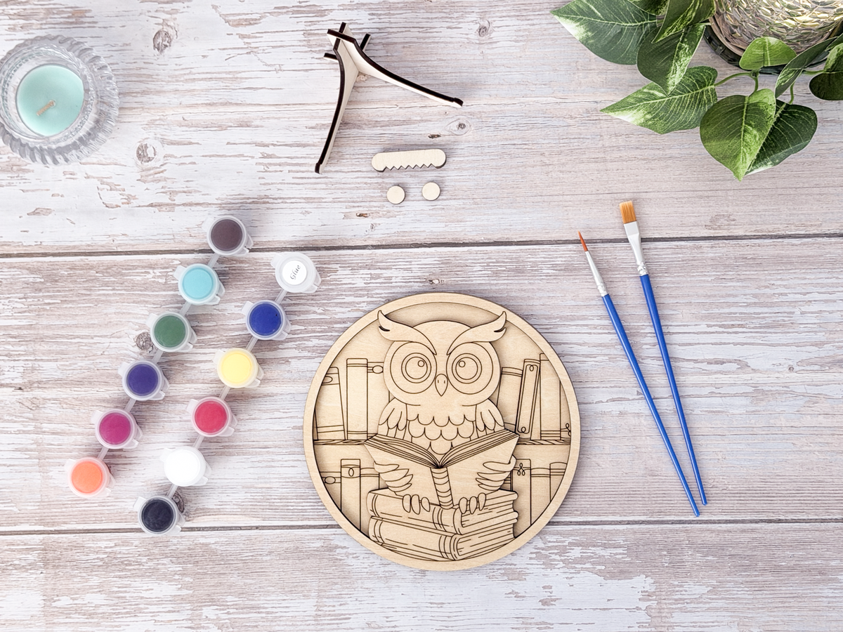 Unpainted book lovers owl layered wood artwork displayed with paint pots, brushes, stand pieces, candle, and greenery.