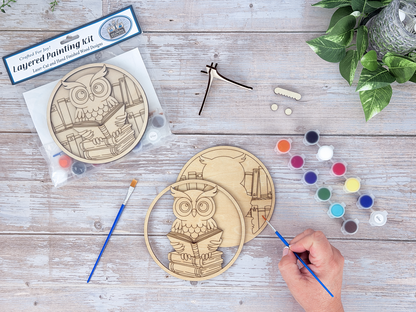 Person painting the book lovers owl layered wood craft kit on a rustic wood background with paint pots, brushes, and greenery.