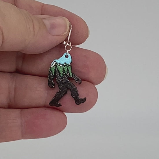Video of an earring shaped like a sasquatch with a stylized mountain scene, showing front and back.