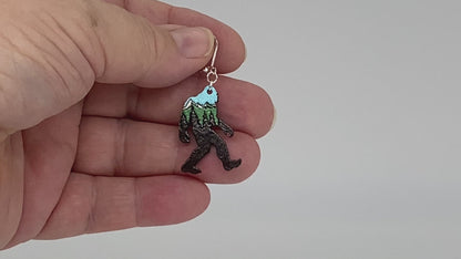 Video of an earring shaped like a sasquatch with a stylized mountain scene, showing front and back.