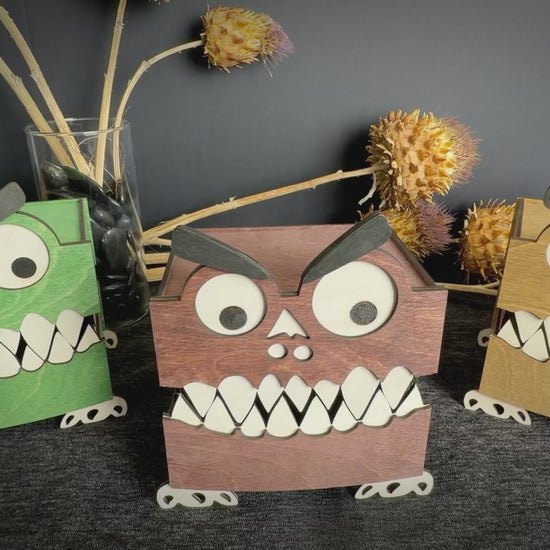 Video of three handcrafted wooden monster boxes in mahogany, green, and walnut stains, featuring googly eyes, white teeth, and playful monster faces.  Hands moving in and displaying middle box showing 360 degrees and inside of box.