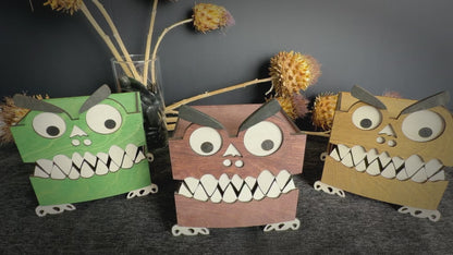 Video of three handcrafted wooden monster boxes in mahogany, green, and walnut stains, featuring googly eyes, white teeth, and playful monster faces.  Hands moving in and displaying middle box showing 360 degrees and inside of box.