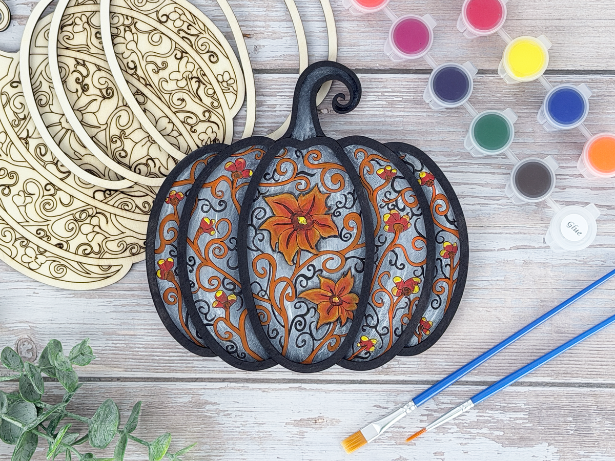 Finished gothic pumpkin adult craft kit shown with painted layered wood artwork, unpainted kit pieces, paint pots, and brushes on a rustic wood background.