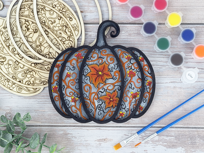 Finished gothic pumpkin adult craft kit shown with painted layered wood artwork, unpainted kit pieces, paint pots, and brushes on a rustic wood background.