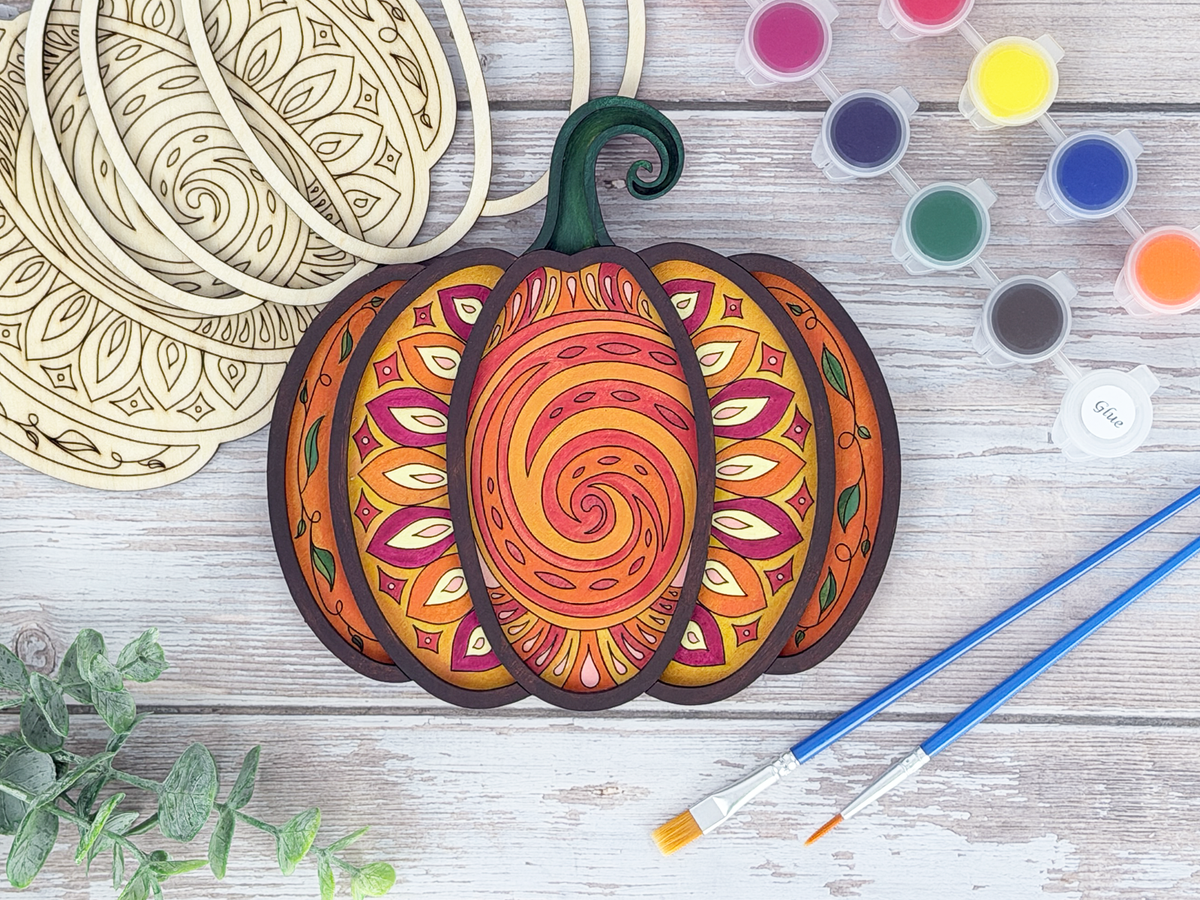 Finished mandala pumpkin adult craft kit shown with painted layered wood artwork, unpainted kit pieces, paint pots, and brushes on a rustic wood background.
